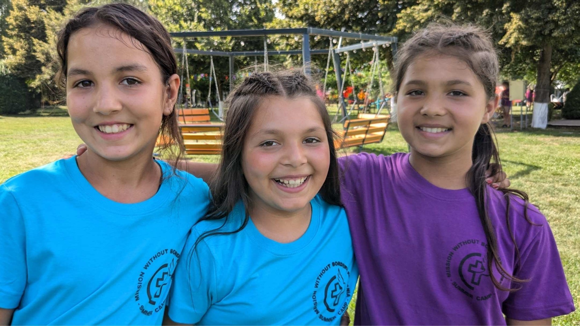 Three girls in camp t-shirts stand close together, smiling broadly with their arms around each other’s shoulders.