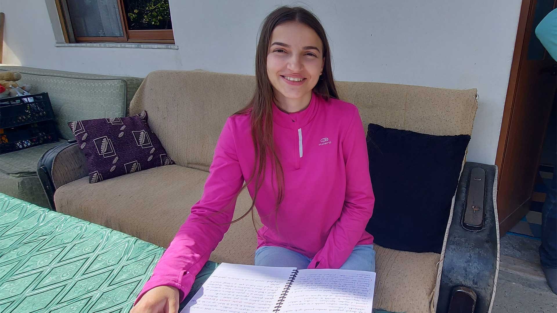 Smiling young woman with dark hair, wearing a pink long-sleeve sweater, sitting on a sofa with an exercise book in front of her.