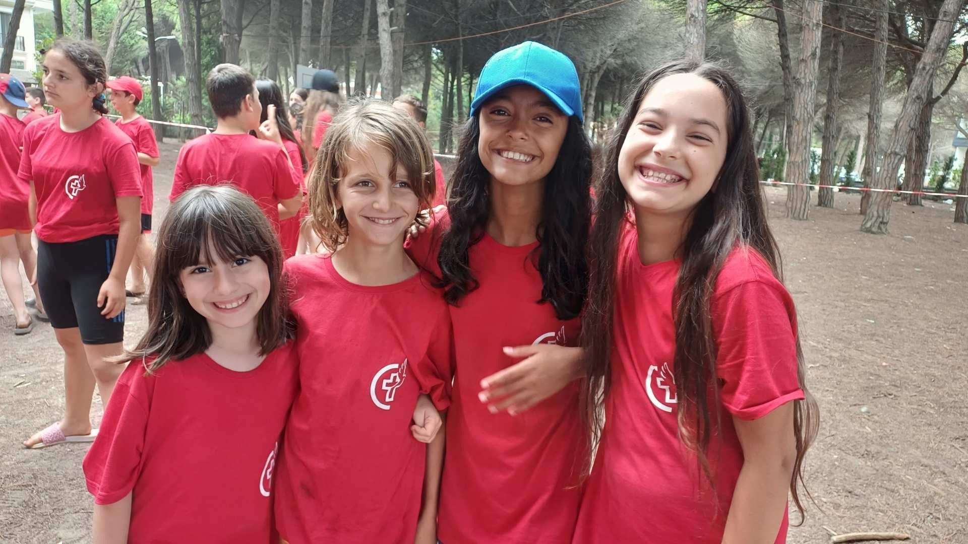 Four young girls in matching summer camp t-shirts standing close together, smiling brightly.