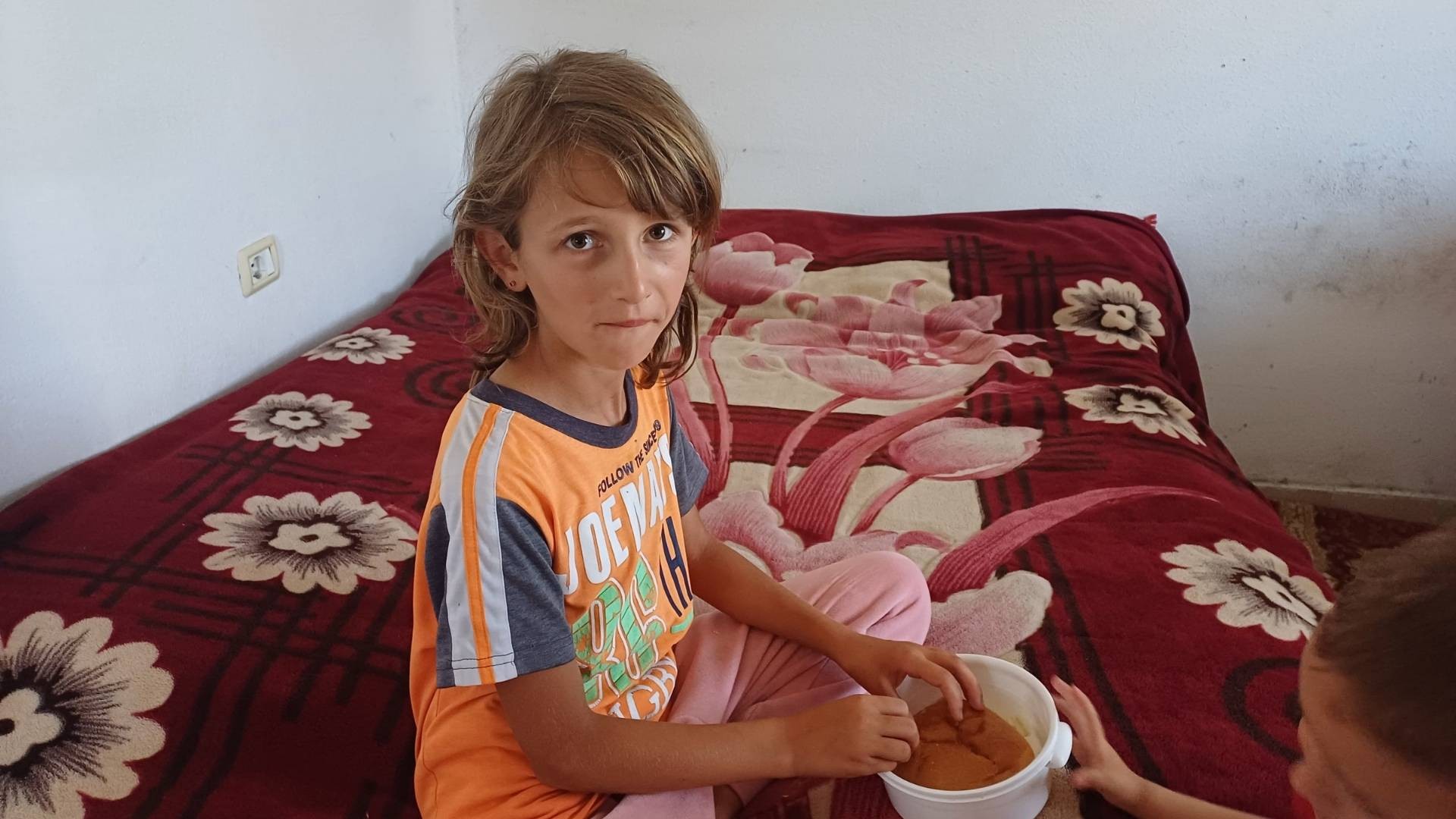 Shy young girl sitting on a bed in a sparsely furnished room, with a bowl of food.