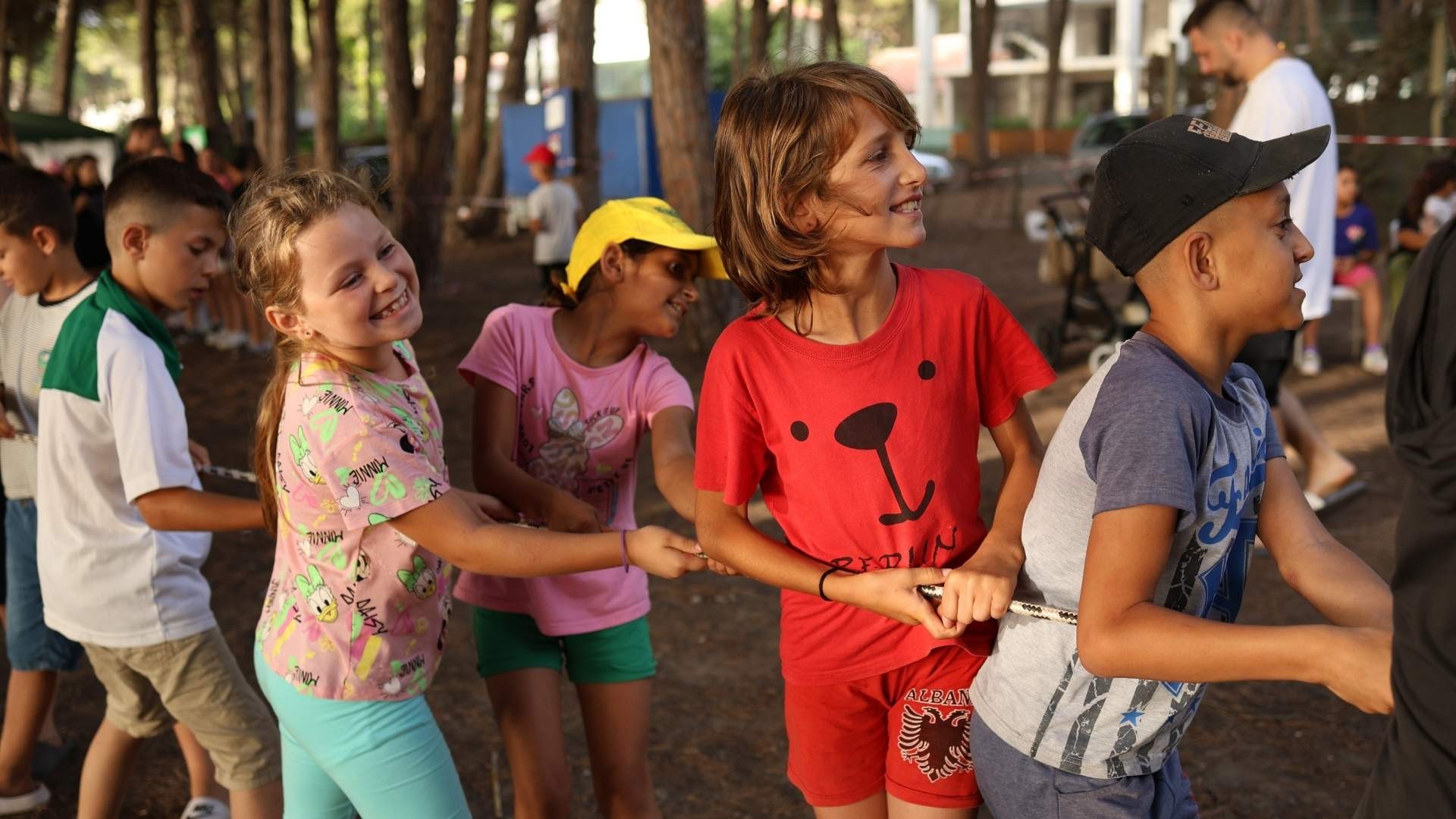 Children playing tug of war under the shade of tall trees in a wooded area.