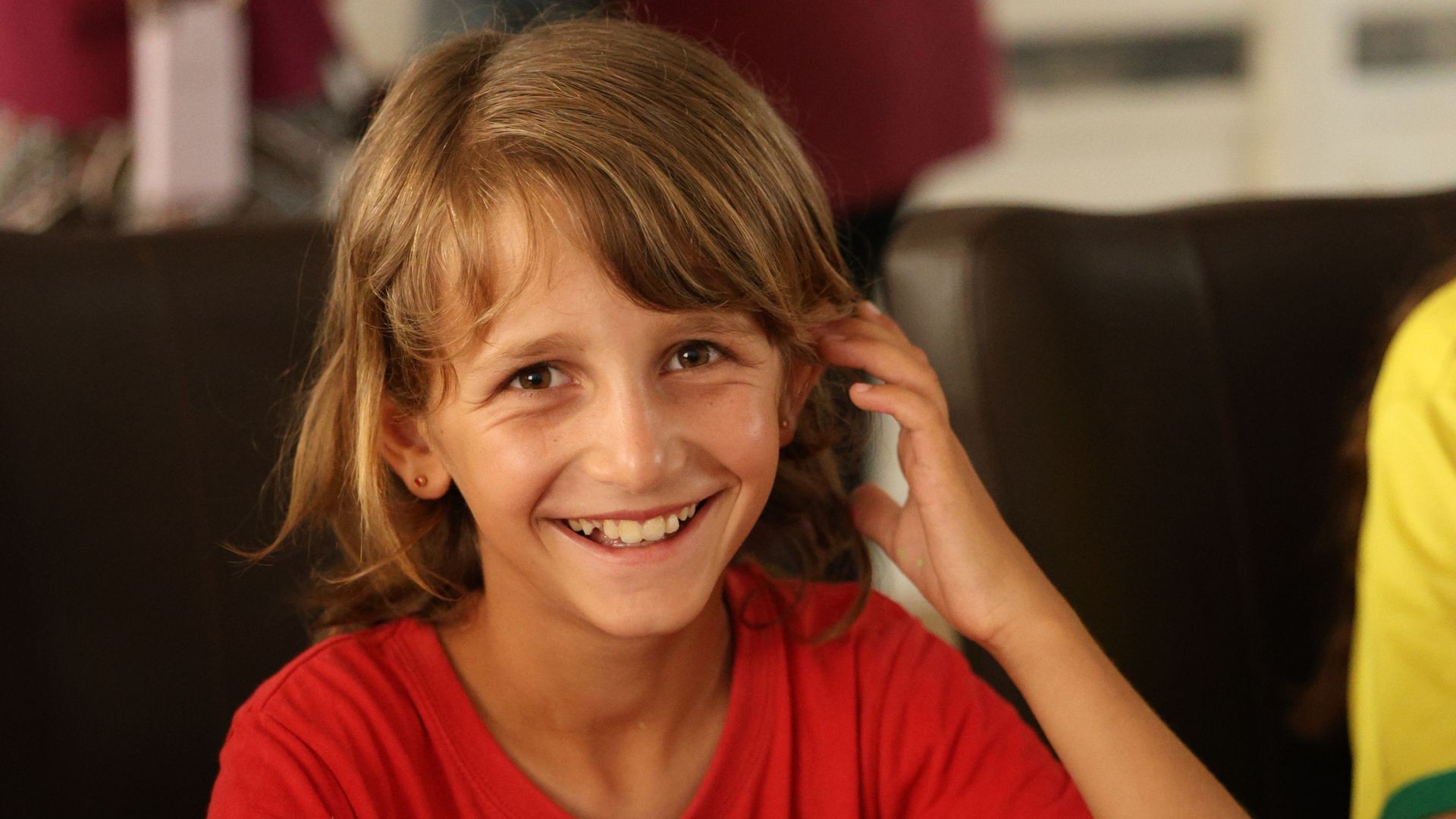 Young blonde girl in red shirt, tucking her hair back with a radiant smile.