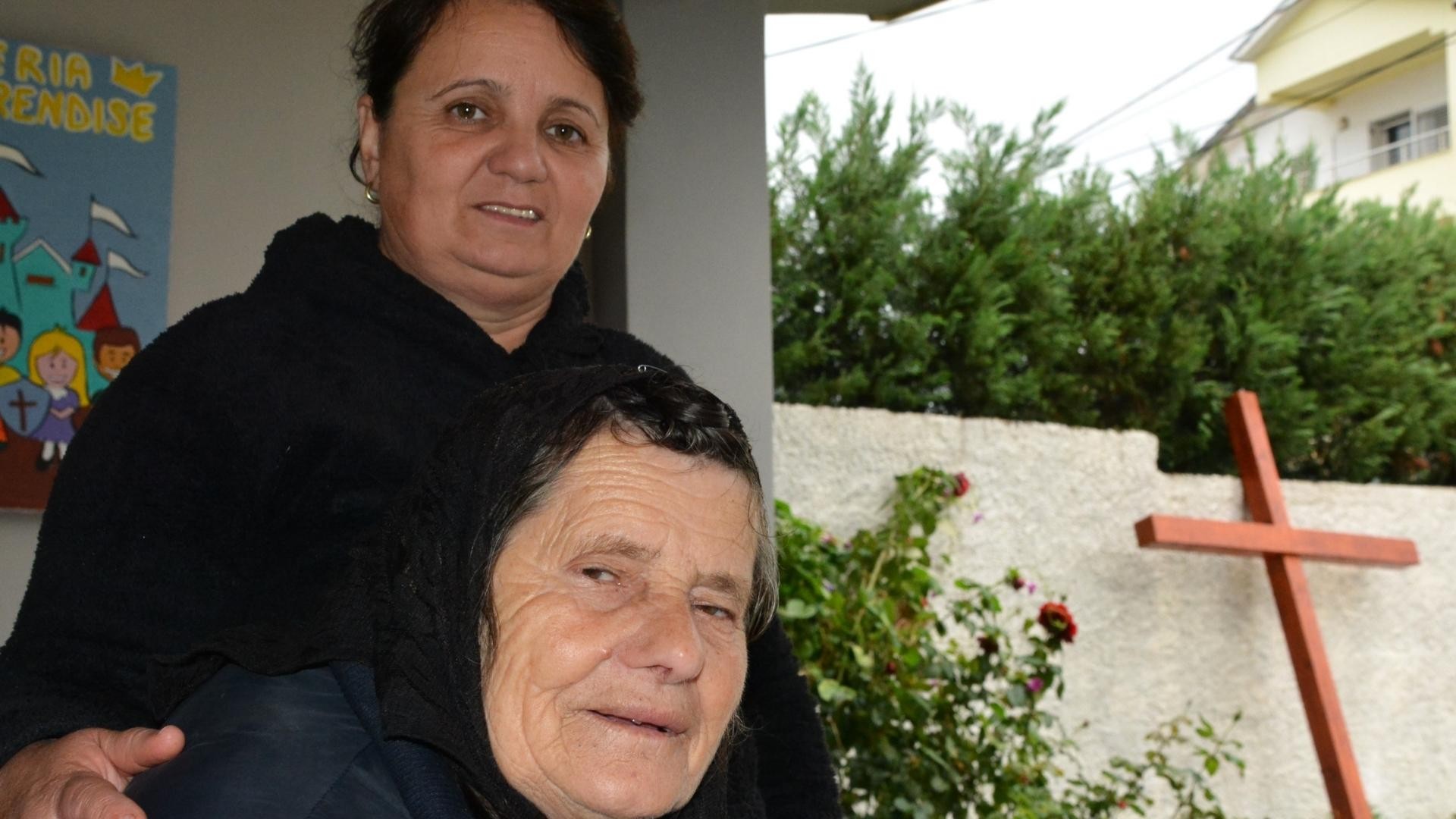 Two women, one seated and the other standing behind her, with a cross visible on an exterior wall in the background.