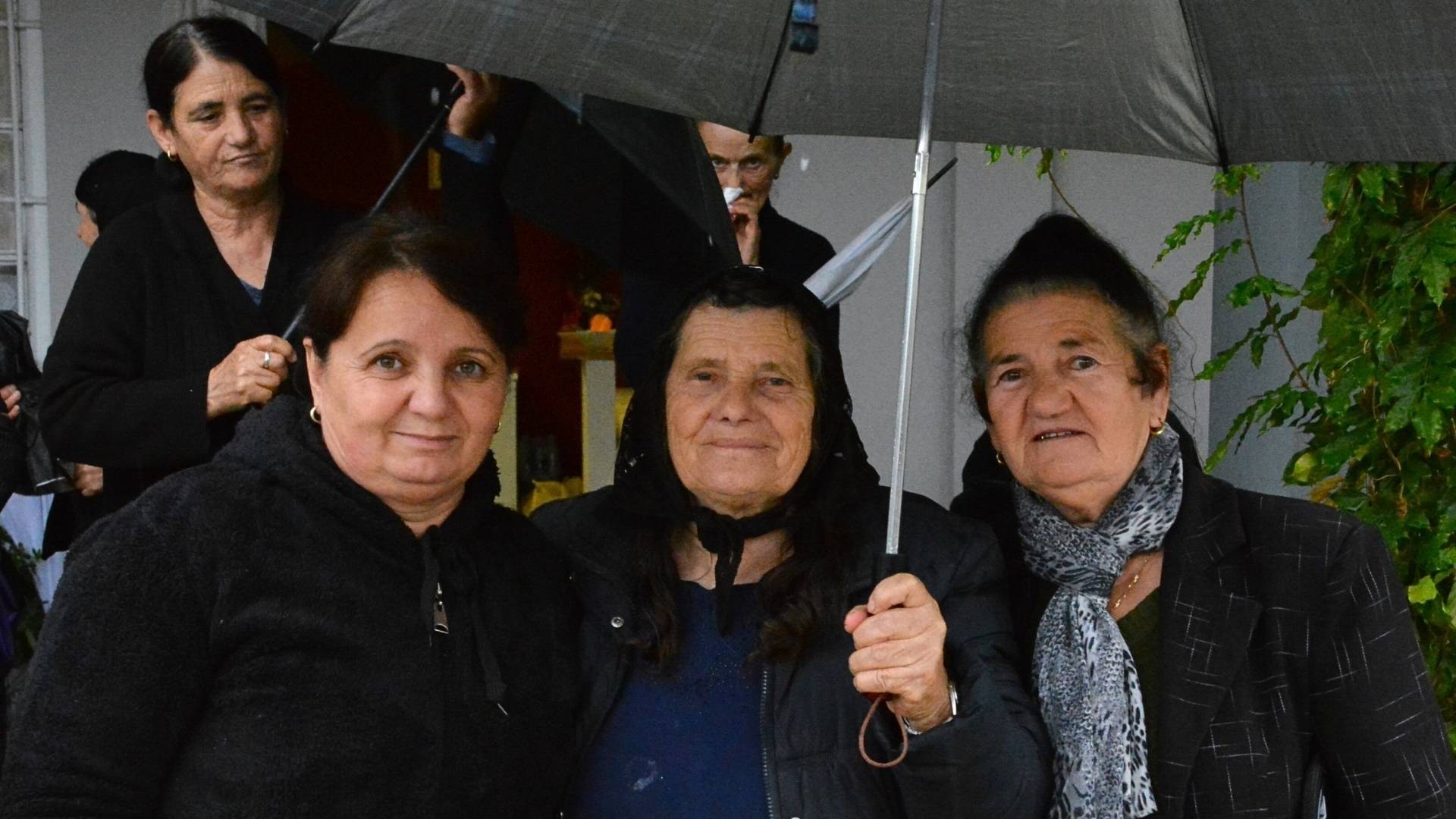 Three women dressed in dark warm clothing standing close together, one holding an umbrella.