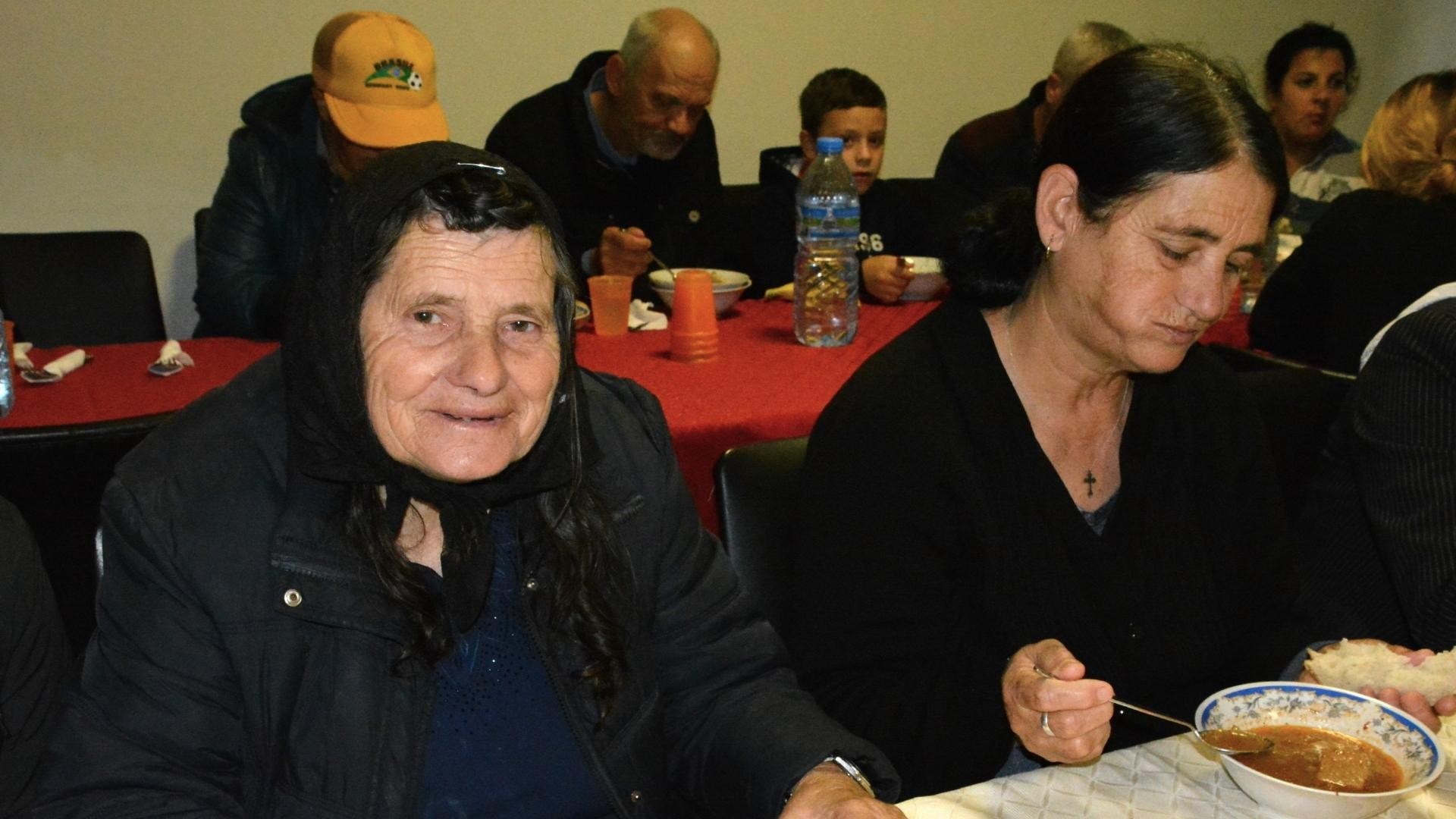 Two elderly women sitting at a table enjoying a warm meal, with other people visible in the background.