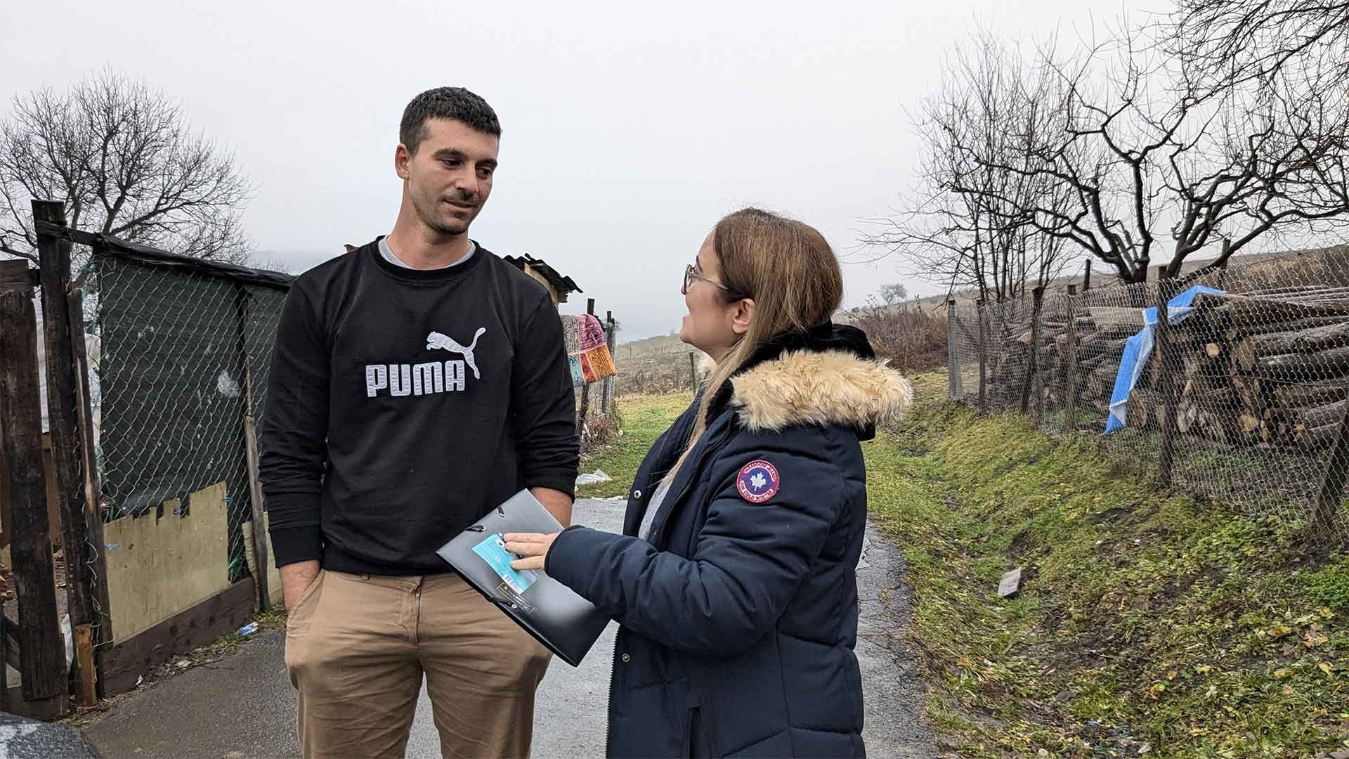 A man and woman talk outside on a winter day, the woman holding a folder as they converse.