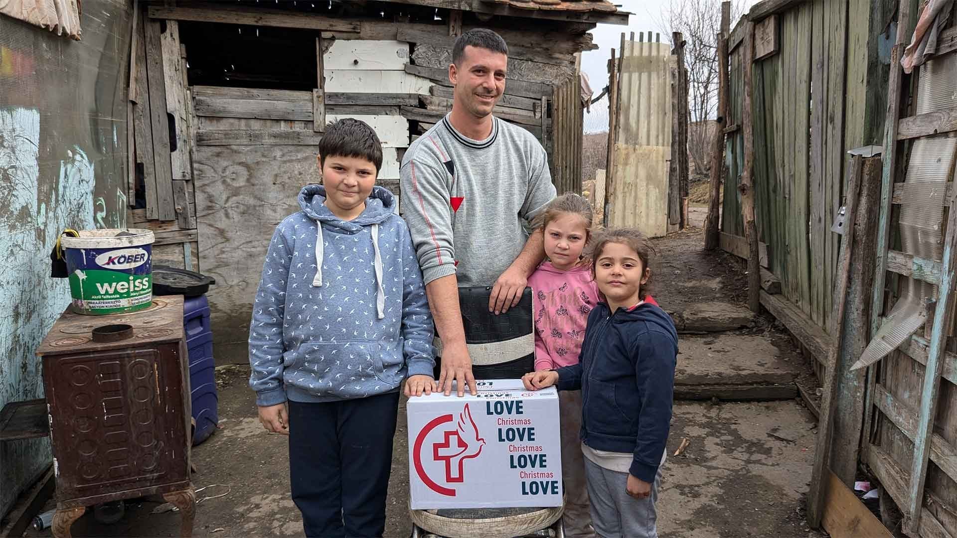 A smiling father and his three children gather around a chair holding a Christmas Love box in their worn yard.