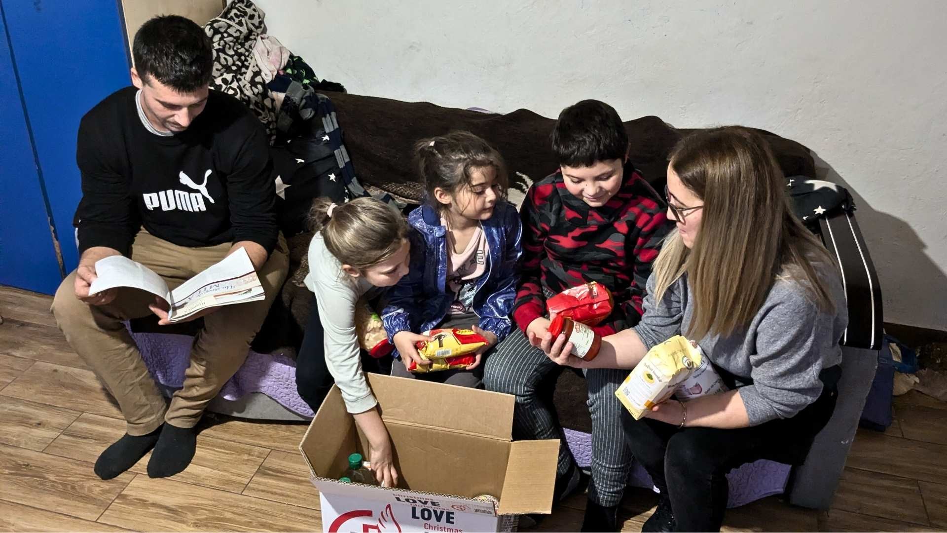 A father and children sit on a couch in a modest room, unpacking a food box with a MWB family worker.