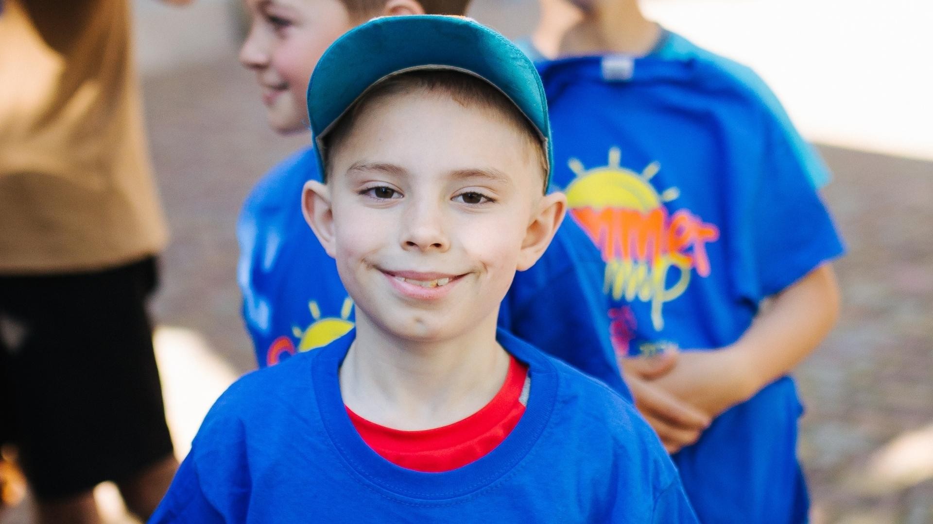 Smiling young boy wearing a blue cap and summer camp t-shirt, with other children in the background.