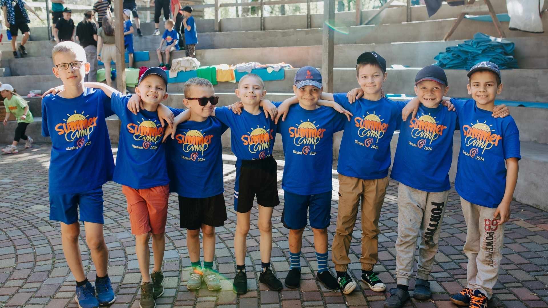Group of young boys wearing matching camp t-shirts, smiling with arms around each other’s shoulders.