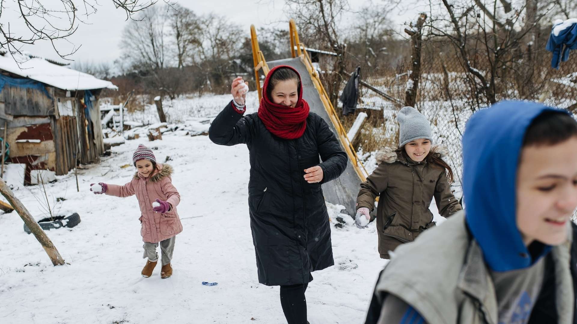 A playful mother laughs with her three children as they run together outside in the snow on a winter day.