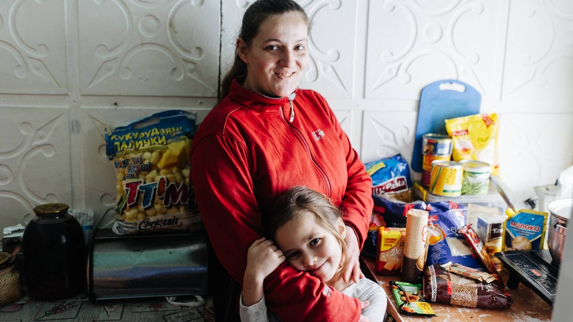 A happy mother hugs her child in their small kitchen as unpacked food items sit nearby on the counter.