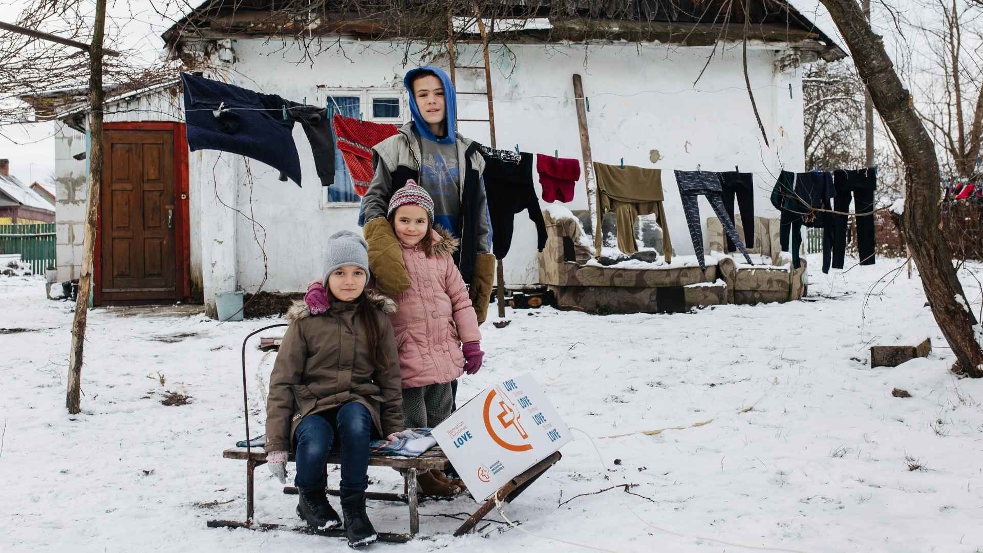 Three children gather around a snow sled with a food parcel, outdoors in front of a clothesline.