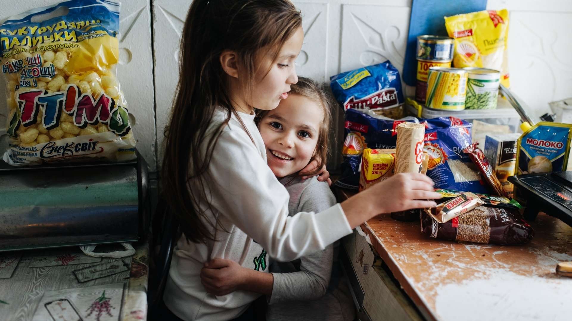 Happy siblings embrace in their small kitchen, surrounded by unpacked food items.