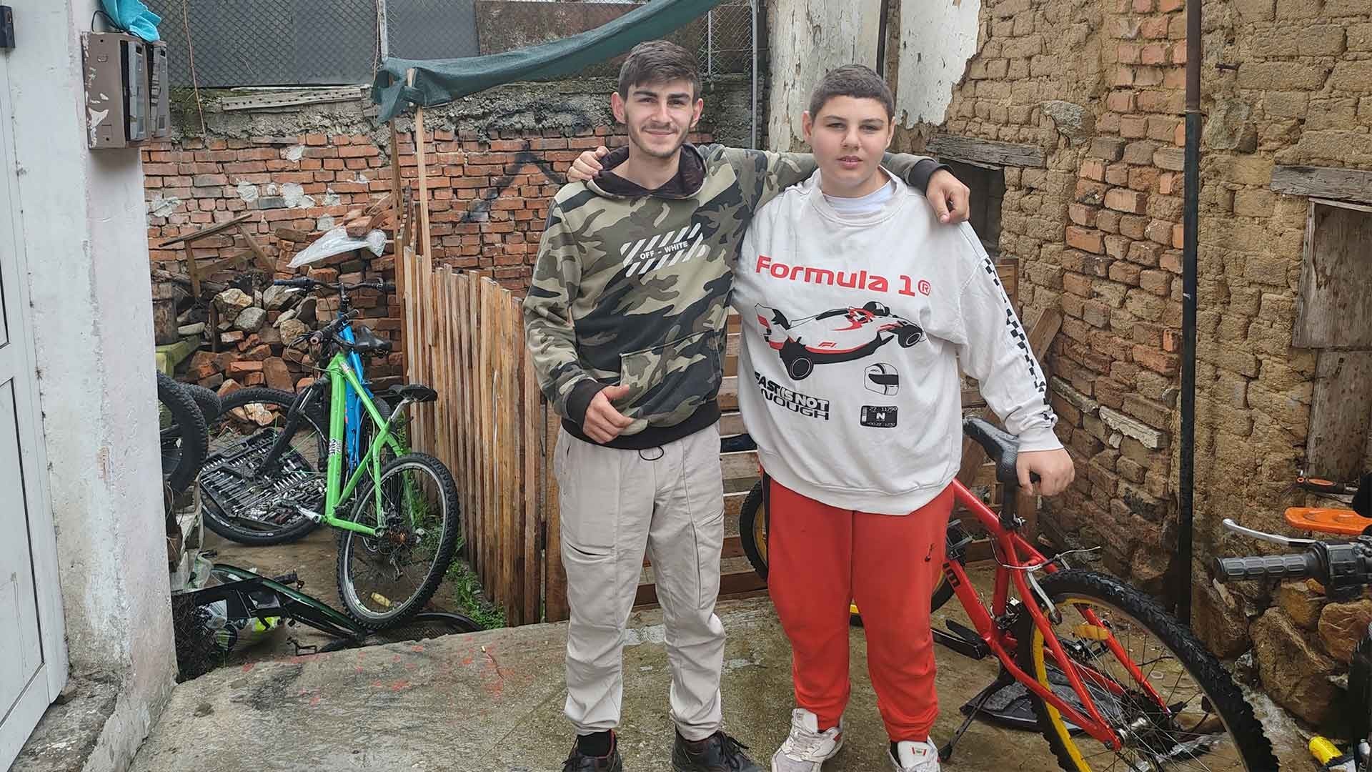 Two young men standing in a backyard with bicycles in the background.