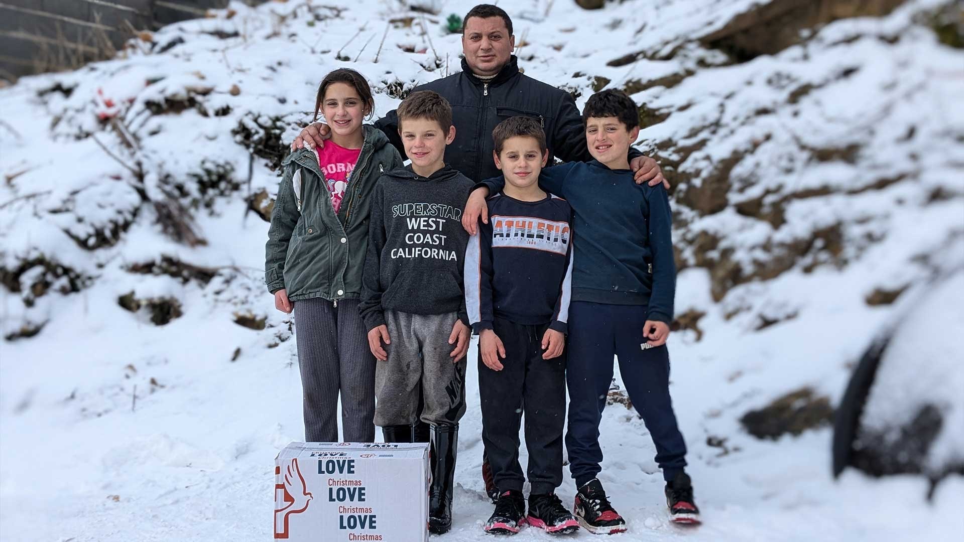 Four children with an older man standing in the snow behind a box on the ground.