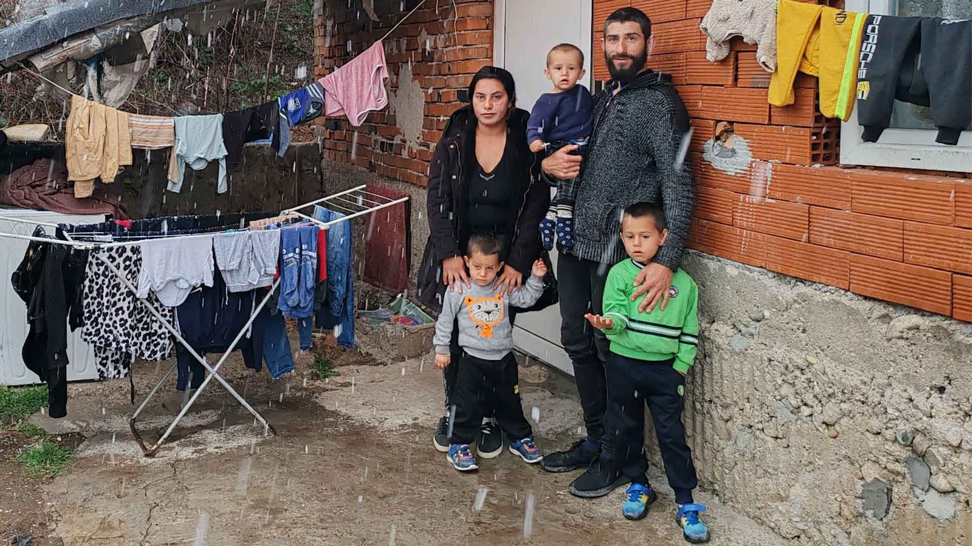 Parents with their three young children standing outside their house as rain falls around them.