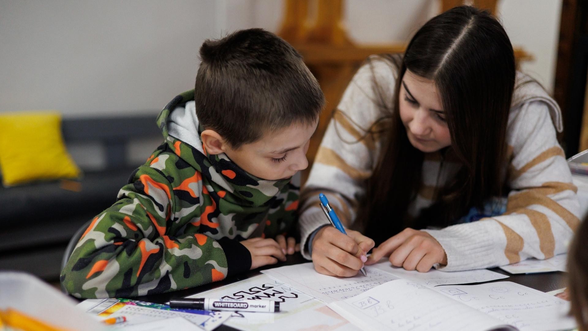 Boy in camouflage sweater concentrates while a young woman shows him how to form alphabet letters