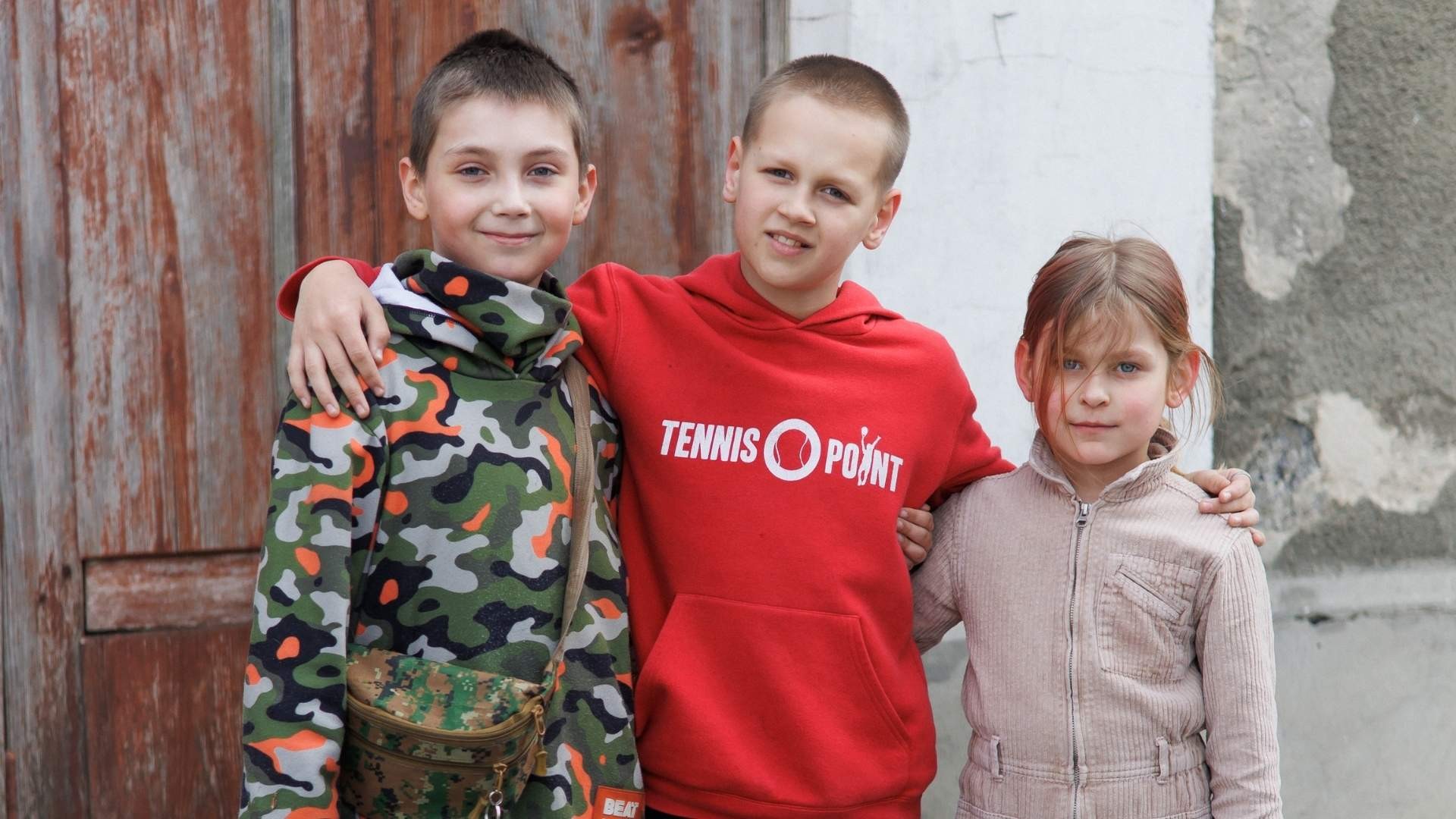 Three young friends stand outside, smiling with their arms around each other’s shoulders.