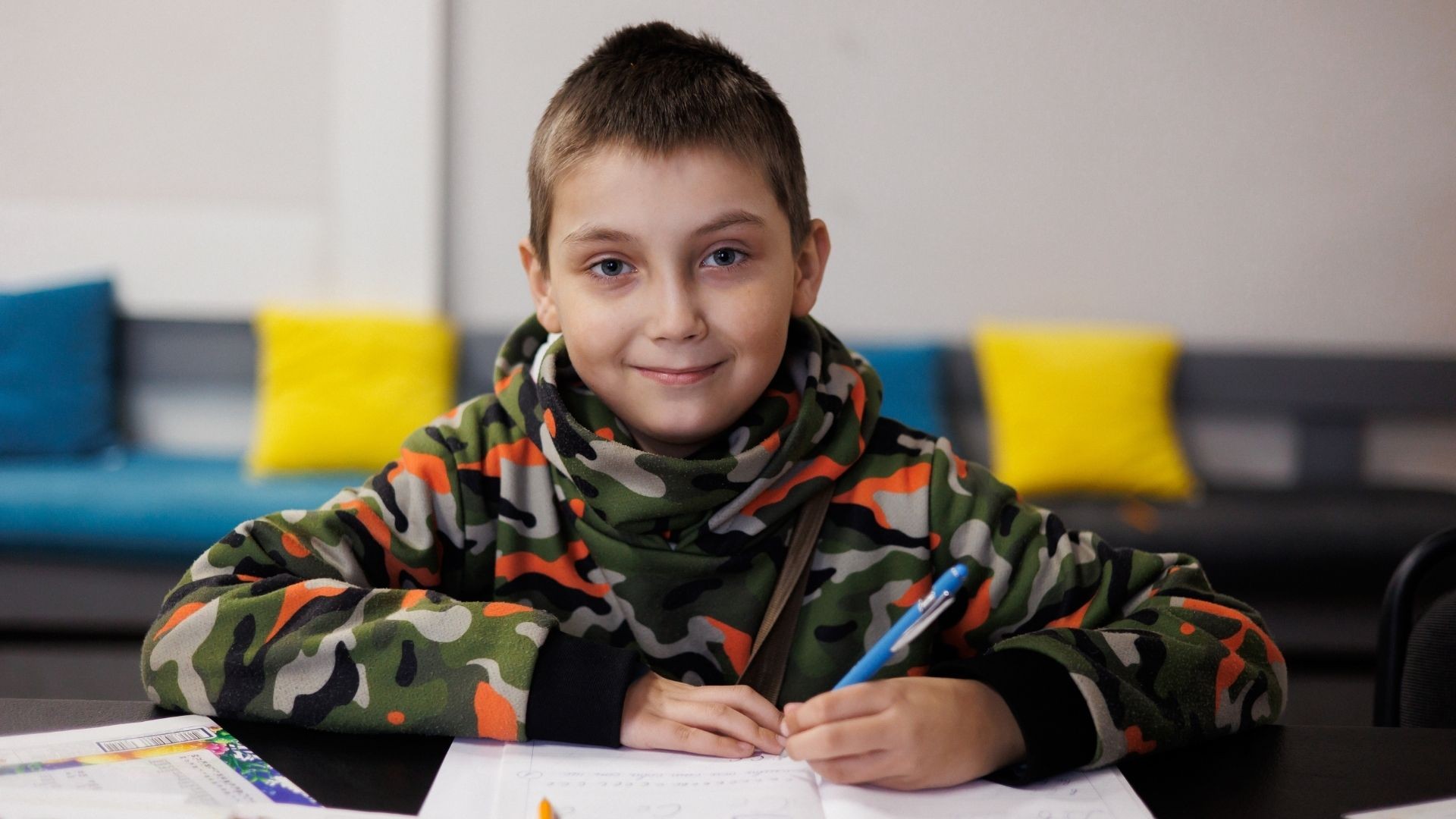 Boy in camouflage sweater smiles as he completes work in his exercise book.