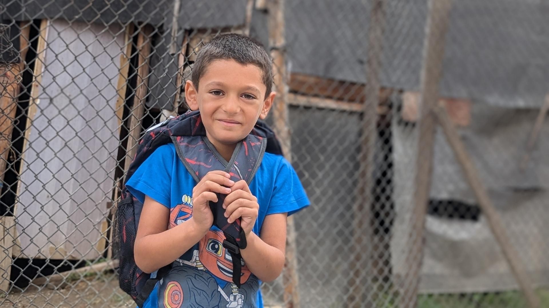 Young Roma boy standing outside a wire fence, smiling shyly as he wears his new backpack.
