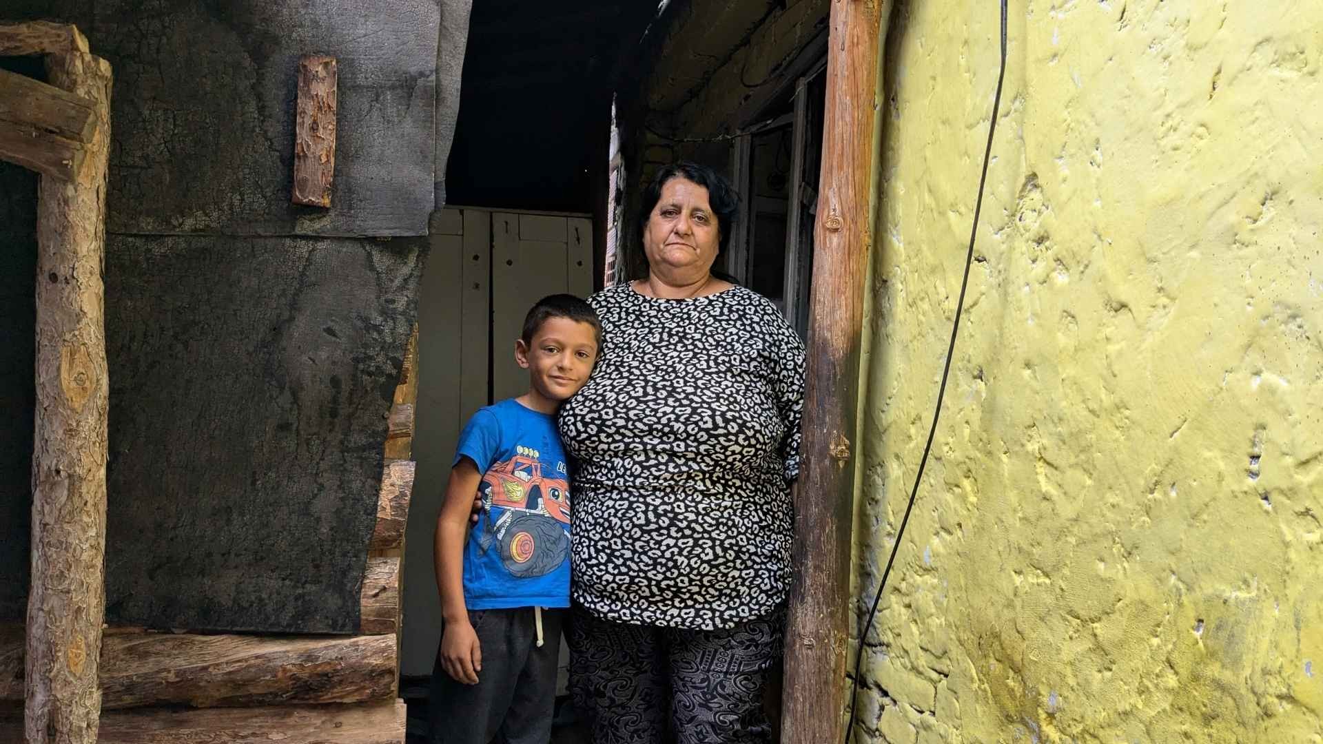 Young Roma boy in a blue t-shirt stands closely embraced by an older Roma woman in a doorway, with a yellow wall on the side.