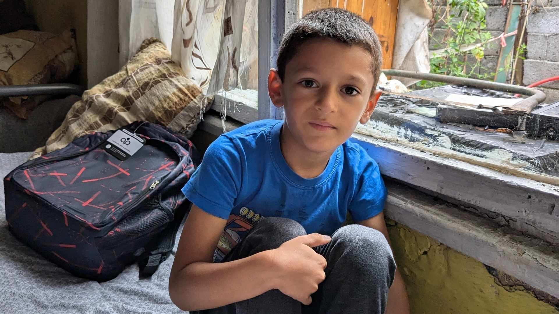 Young Roma boy in a blue t-shirt sits on a bed beside an open window, with a new backpack placed behind him.