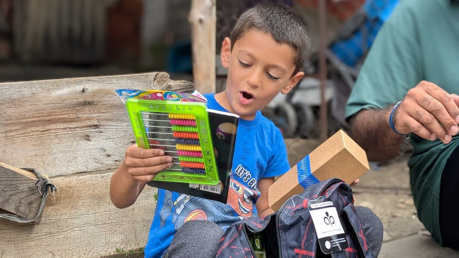 Young Roma boy sitting on a step outside, excitedly looking through new school supplies he’s taken from a new backpack, with someone seated beside him.